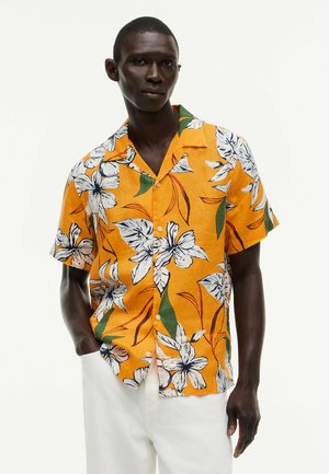 Mężczyzna o ciemnej karnacji w jaskrawo pomarańczowej koszuli z krótkim rękawem z dużymi białymi kwiatowymi wzorami oraz białych spodniach na jednolitym tle.