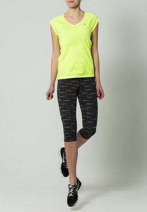 Vrouw draagt een neongele T-shirt met korte mouwen, zwart-witte capri leggings met patroon en zwarte hardloopschoenen tegen een effen achtergrond.