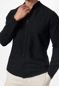 Czarna koszula z długim rękawem wykonana z miękkiej tkaniny, z kołnierzykiem zapinanym na guziki, kieszenią z przodu oraz sześcioma guzikami na przedniej listwie.