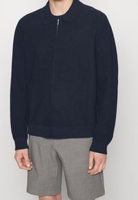 Námořnická žebrovaná bunda na zip s límcem, která má dvě boční kapsy a přiléhavý pas, spárovaná se šedými kostkovanými šortkami.