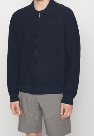 Veste zippée en côtelé marine avec col, dotée de deux poches latérales et d'une taille ajustée, associée à un short gris à carreaux.