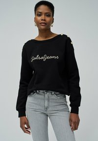 Sweatshirt noir avec encolure bateau, accents de boutons dorés sur les épaules, et "SalsaJeans" brodé en argent. Associé avec un jean gris clair.