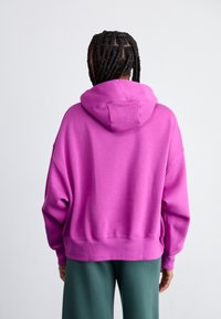 Persona con cabello trenzado que lleva un hoodie púrpura brillante y pantalones verde oscuro, de espaldas contra un fondo claro.