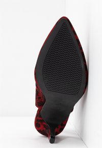 Escarpin à talon haut imprimé léopard rouge à bout pointu, vu de la semelle, avec une semelle en caoutchouc noir texturé, appuyé contre un mur blanc.