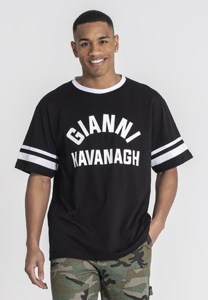 Camiseta negra de gran tamaño con texto blanco "GIANNI KAVANAGH" y rayas blancas en las mangas. Combinada con pantalones de camuflaje.