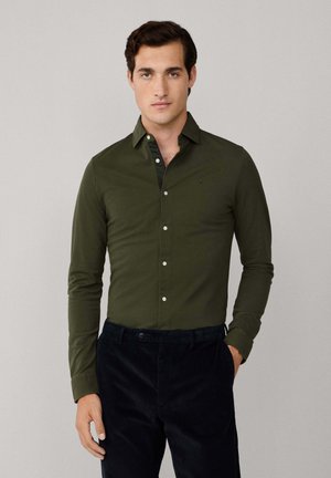 Olive green button-up shirt with a pointed collar, long sleeves, and white buttons. Worn with black corduroy pants. Textured fabric.