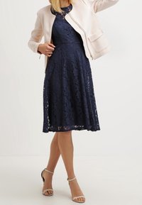 Navy lace dress with a flared skirt, complemented by a light pink, structured jacket. Features fitted bodice and subtle detailing. Silver sandals visible.