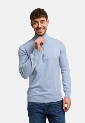Smilende mann med kort krøllete hår iført en lyseblå genser med glidelås og mørkeblå jeans, justerer genserens krage.