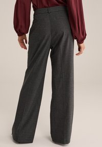 Persona che indossa pantaloni a quadri grigi a vita alta e gamba larga, accompagnati da una blusa lunga borgogna, in piedi con le mani rilassate.