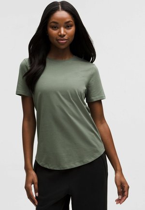 Olijfgroene T-shirt met korte mouwen, ronde hals en een afgeronde zoom. Zachte, lichte stof met een gladde textuur. Gecombineerd met een zwarte broek.