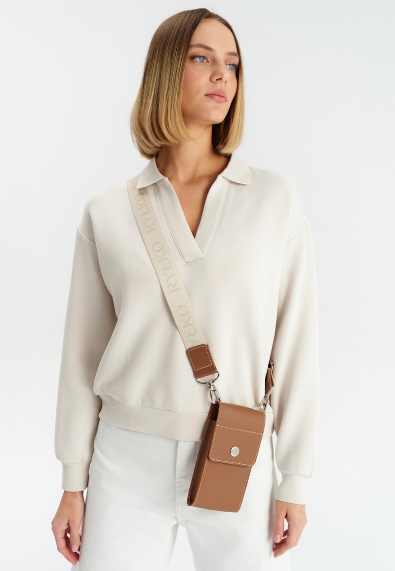 Brun læder crossbody taske med en tekstureret overflade, sølv hardware og en rem med et logo. Bæres over en lys langærmet top.