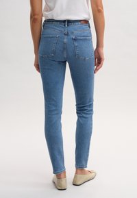 Jeans ajustados de color azul con bolsillos traseros de parche, denim ligeramente desgastado, cintura alta y detalles de costura mínimos, combinados con zapatos planos beige.