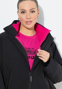Kvinna i en svart jacka med en ljusrosa innerfoder och en matchande rosa t-shirt med grafik, som delvis öppnar jackan.
