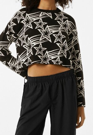 Pull noir court avec motifs d'étoiles filantes blanches porté avec un pantalon noir ample, la main du modèle posée sur la hanche.