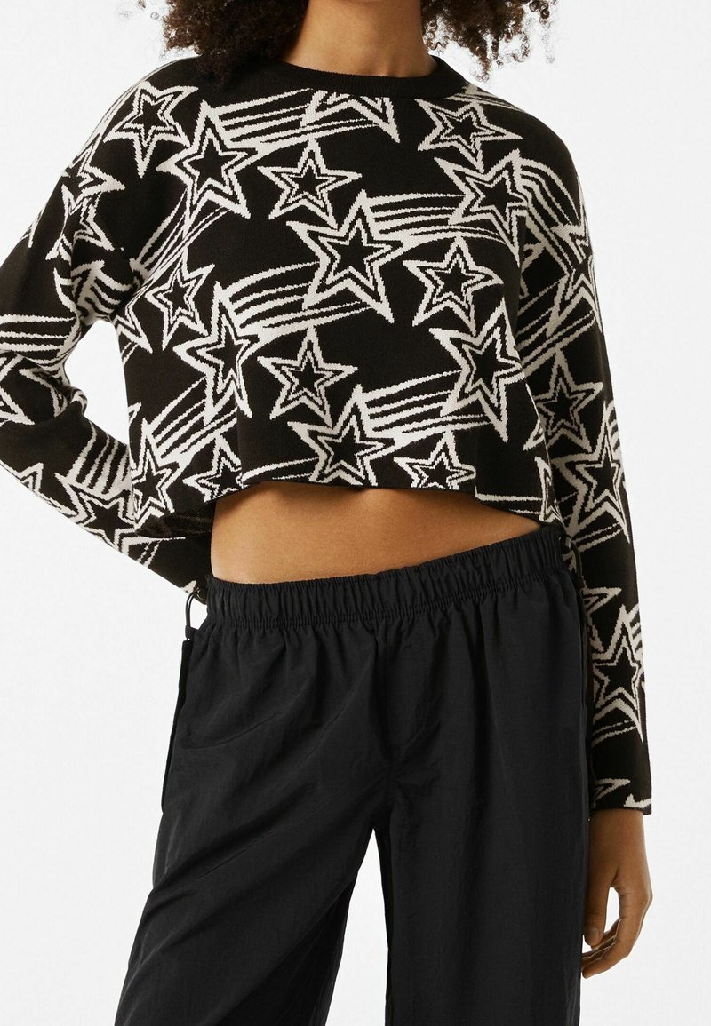 Pull noir court avec motifs d'étoiles filantes blanches porté avec un pantalon noir ample, la main du modèle posée sur la hanche.