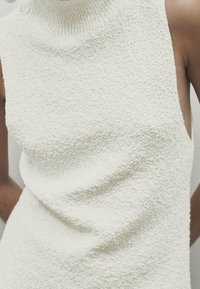 Haut sans manches couleur crème avec un col montant, fabriqué en tissu bouclé texturé, présentant une silhouette ajustée et des côtés ouverts.