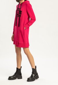 Kobieta ubrana w jasnoróżową sukienkę z kapturem z czarnym napisem i czarnymi, masywnymi botkami na kostkach, stoi z jedną ręką w kieszeni na białym tle.