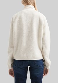 Veste en polaire de couleur blanc cassé avec un extérieur texturé et pelucheux. Présente un col montant et une coupe décontractée, associée à un jean bleu foncé.