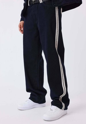 Personne portant un pantalon noir avec des bandes latérales en dentelle blanche et des baskets blanches, debout devant un fond clair uni.