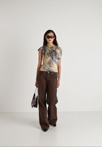 Bruine wijde broek met een geel en grijs patroon bovenstuk. Accessoires zijn een zwarte handtas en donkere zonnebril. Glad textuur.