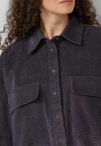 Korduroy shirt in donkerpaars met knoopsluiting aan de voorkant, puntige kraag en twee zijzakken. Heeft een verticale ribbelstructuur.