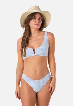 Vrouw die een lichtblauw-wit gestreepte bikini en een beige hoed met brede rand draagt, poserend tegen een egale lichte achtergrond.