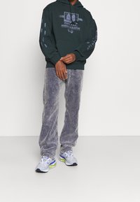 Tmavě zelená mikina s kapucí s grafickou potiskem, žebrovanými manžetami a klokankovou kapsou. Kombinována se světle šedými manšestráky a mnoho barevnými teniskami.