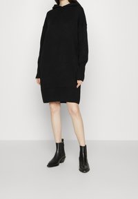 Vestito a maglione oversize con cappuccio nero in morbido tessuto, con polsini e orlo a coste, caratterizzato da una scollatura a V. Abbinato a stivaletti neri alla caviglia.