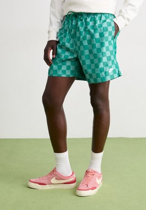 Person iført grønternet shorts med Nike-logo, hvide sokker og pink Nike-sneakers, stående på grøn gulv med hænderne i lommerne.