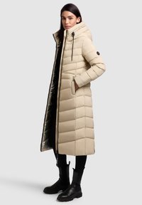 Hosszú bézs színű steppelt kabát varrott panelekkel, állógallérral, elülső zsebekkel és cipzáras záródással. Belseje sötétebb kontrasztos béléssel rendelkezik.