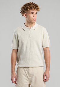 Lühikeste varrukatega beež polo särk, millel on sooniktextuur ja neljanda osa tõmbluku kaelus. Omab lõdva lõikega ja tagasihoidlikke külgaktsente.