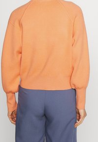 Suéter naranja de punto con mangas abullonadas, puños acanalados y un dobladillo corto, combinado con pantalones azules holgados. Textura suave evidente.