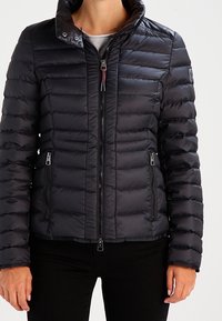 Veste matelassée noire avec fermeture éclair, col montant et design matelassé horizontal. Comprend deux poches avant avec fermetures éclair.