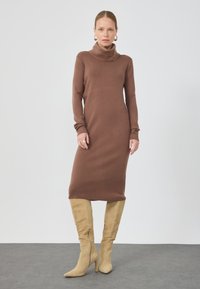 Rochie lungă tricotată, maro, cu mâneci lungi și gât înalt, cu o siluetă ajustată. Purtați-o cu cizme bej, până la genunchi, cu vârf ascuțit, din suede.