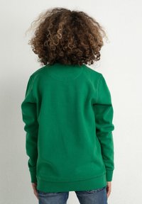 Barn med lockigt hår som bär en grön sweatshirts och blå jeans, står vänd mot en enkel vit vägg.