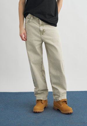 Személy laza, világos bézs farmert, barna munkavédelmi cipőt és fekete pólót viselve, kék szőnyeggel borított padlón áll.