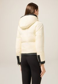 Puffer jacket in light cream with a black hood and cuffs. Features a fitted design and zippered accents on the sleeves. Soft, shiny fabric.
