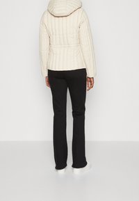 Veste matelassée beige clair avec capuche, silhouette cintrée et taille ceinturée. Associée à un pantalon noir et des chaussures à plateforme blanches.