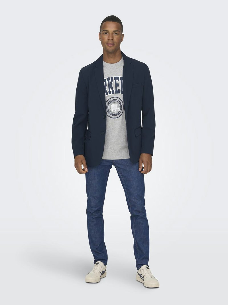 Blazer blu navy sopra una maglietta grigia con stampa "Parker", abbinato a jeans blu scuro e sneaker bianche, in piedi su uno sfondo neutro.
