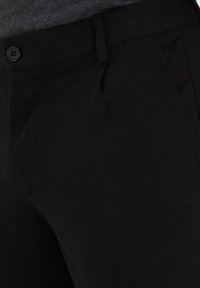 Svarta byxor i texturerad väv, med en framknäppning och subtila veck, som visar på skräddarsydd design och rena linjer.