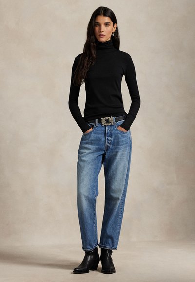 Naine, kes kannab musta kõrge kaelusega sviitrit, kõrge värvliga siniseid teksapükse dekoratiivse vööga, musti pahkluudseid saapaid, seisab käed taskutes.