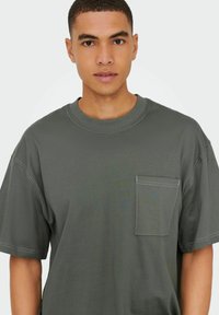 Grå bomulds t-shirt med rund halsudskæring, korte ærmer og en lomme på venstre bryst. Har kontrasterende syninger og en afslappet pasform.