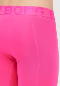 Jasne ružové legíny s hladkou textúrou, s širokým pásom embosovaným s názvom značky "BORG." Viditeľný šev na nohe.