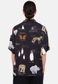 Camisa negra de manga corta con un patrón de varios animales, incluyendo pingüinos, tigres y una mariposa, con nombres científicos impresos.