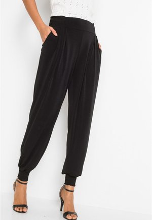 Femme portant un pantalon noir taille haute, coupe fuselée, avec des poignets élastiques et des poches latérales, associé à des sandales noires à talons hauts.