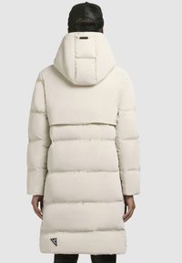 Cappotto lungo e voluminoso di un beige chiaro, dotato di cappuccio, con motivo trapuntato orizzontale e un'acca di logo sul lato inferiore. Texture liscia.