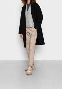 Czarny długi płaszcz, szary sweter z guzikami, szara koszulka, beżowe spodnie, kremowe sneakersy; gładki materiał, luźny krój, warstwowy styl.