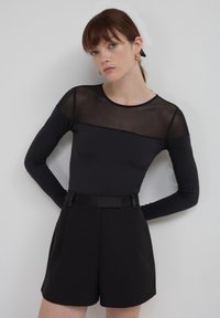 Top nero a maniche lunghe con pannello trasparente sulla parte superiore, abbinato a pantaloncini neri a vita alta con cintura. Tessuto morbido, vestibilità moderna.