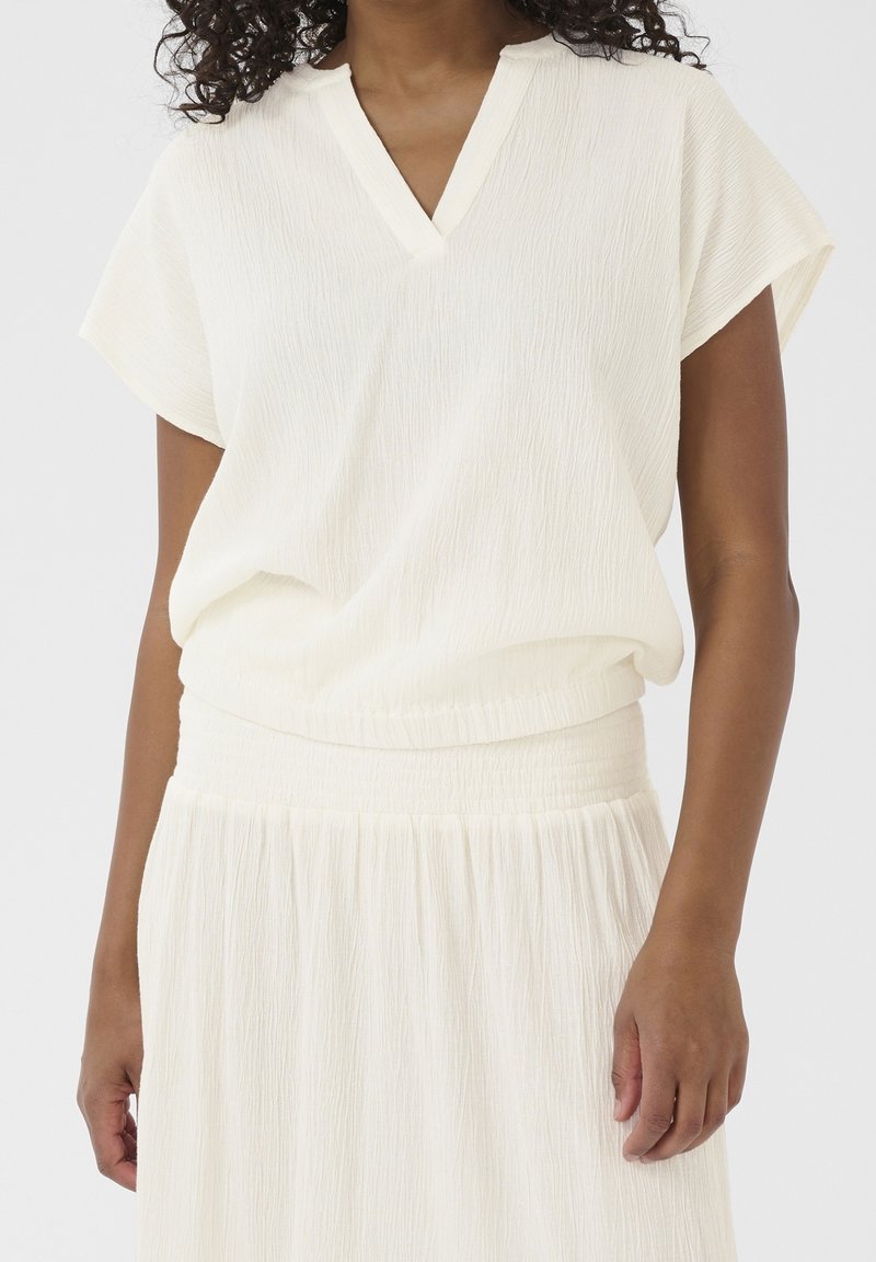 Femme portant un chemisier blanc texturé à manches courtes avec un col en V et une jupe blanche texturée assortie sur un fond uni.