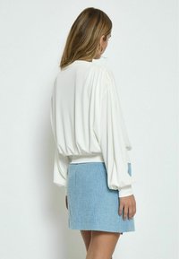 Weißes Longsleeve-Oberteil mit gerafften Schultern, kombiniert mit einem strukturierten blauen Minirock mit vertikalen Streifen. Schlicher Hintergrund.
